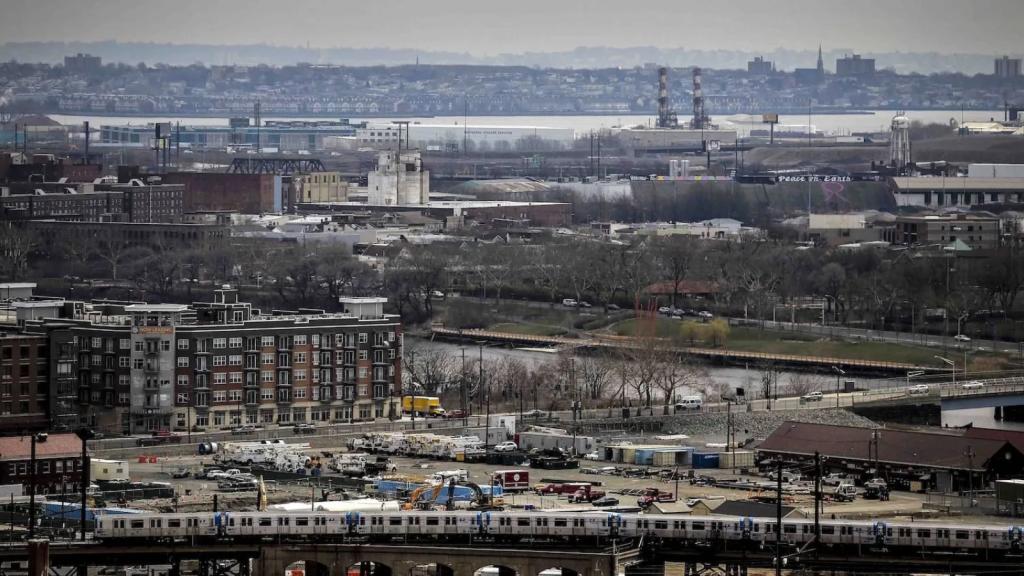 train passes in front of apartment buildings with industrial facilities and a river in the background