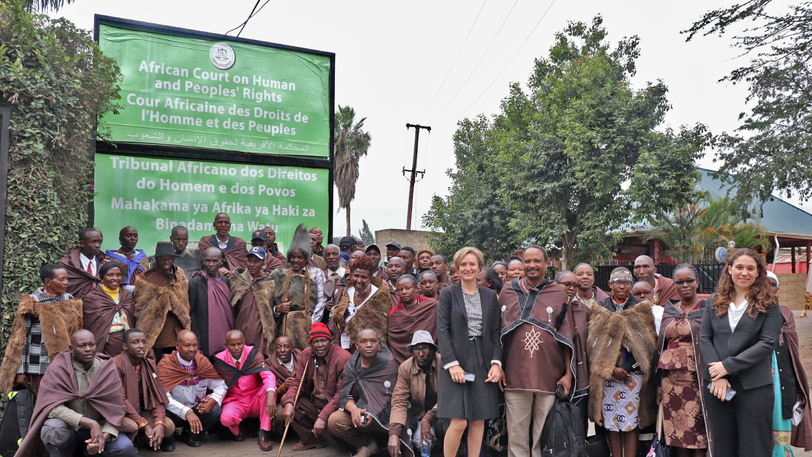 A group of Ogiek people, an indigenous tribe native to Tanzania and Southern Kenya, posing in front of a green sign for the African Court on Human and Peoples' Rights