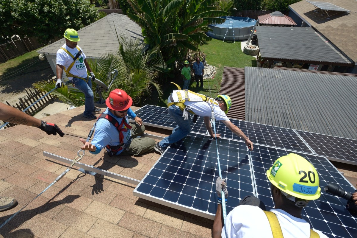 A photo of four men in hard hats on a rooftop, installing blue solar panels. They are bound by safety harnesses. Two men watch them from the ground.