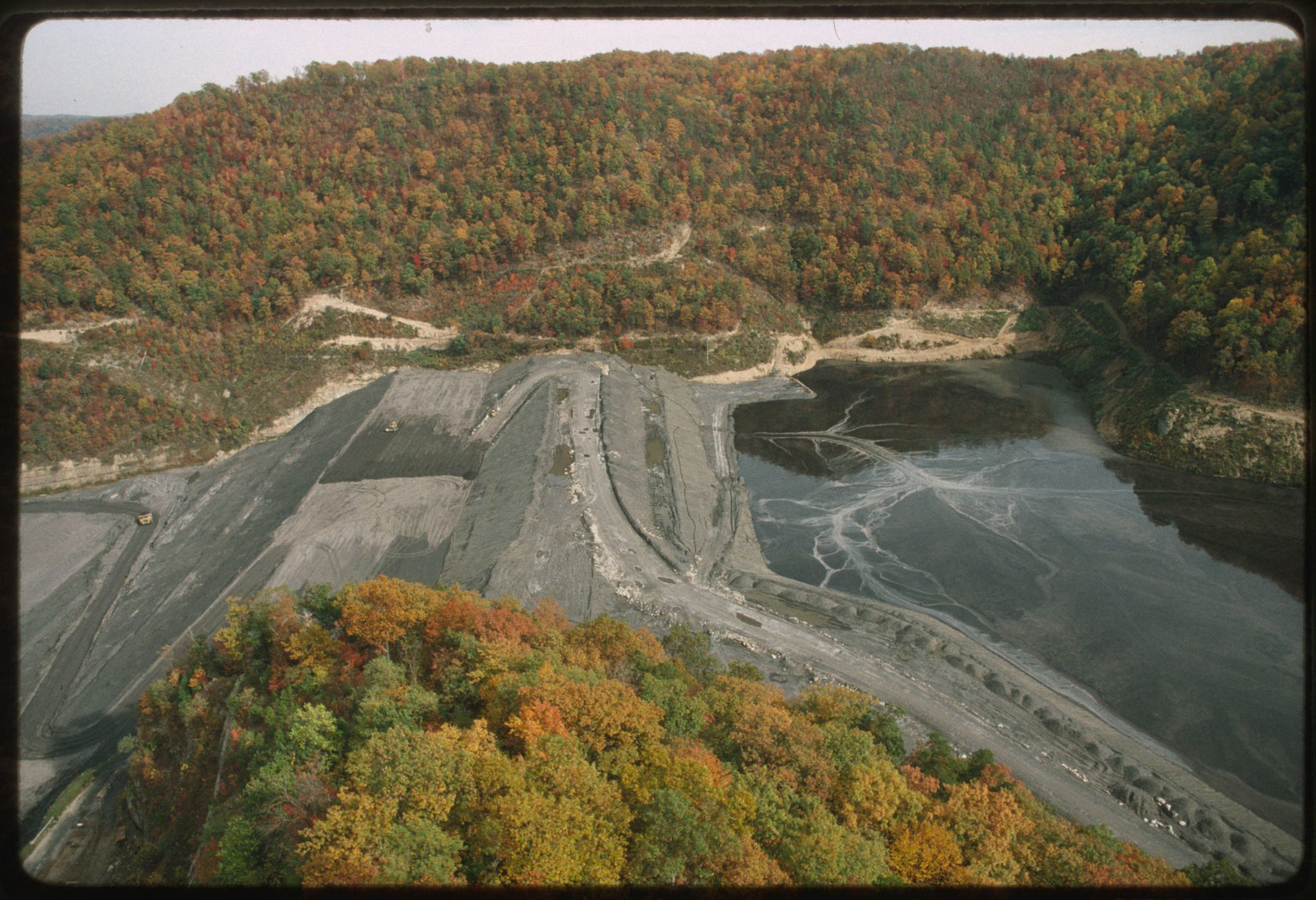 Aerial view of a coal mining "sludge dam"; tree covered mountains surround a cleared area that leads to a dam filled with gray sludge