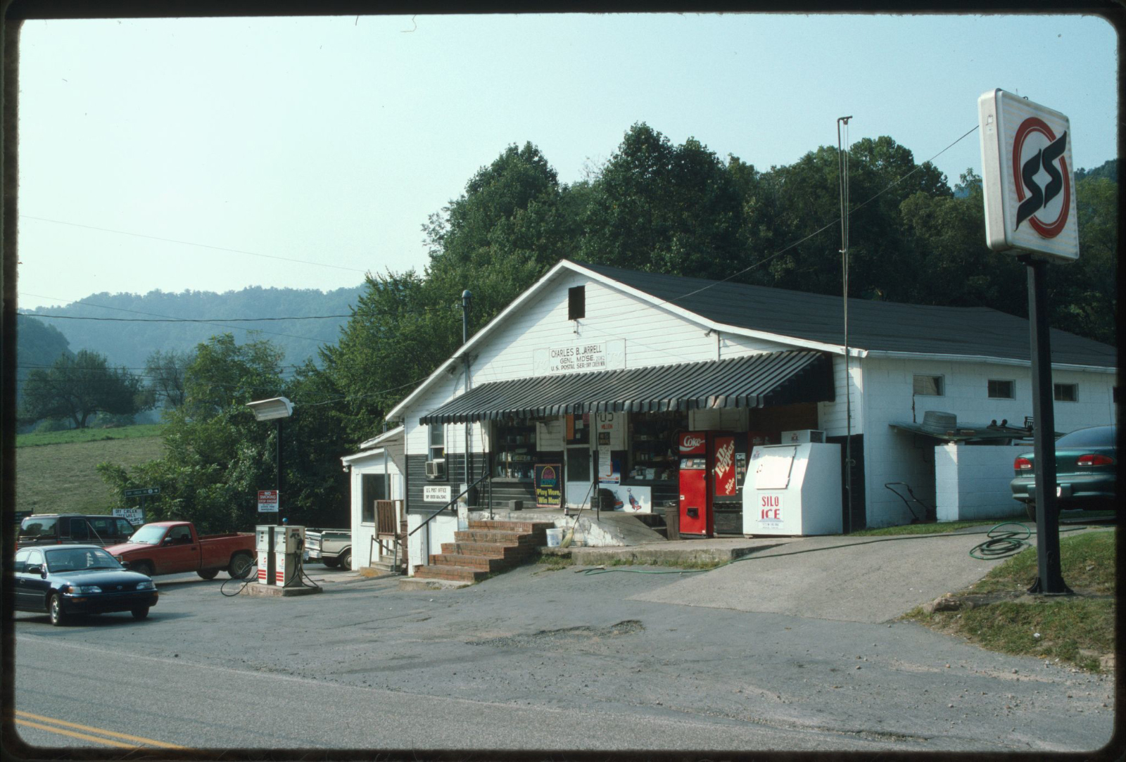 Small white convenience store with black awning and ice and soda machines out front, with a small gas pump to the left and trees in the background