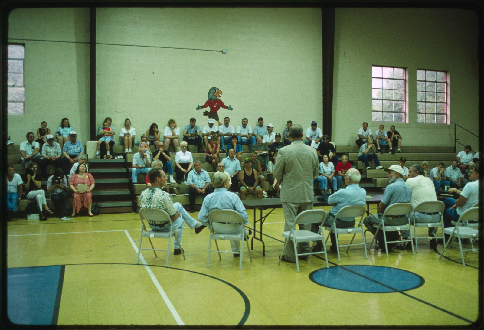 Small group of people on folding chairs addressing a crowd sitting in the bleachers of a school gymnasium