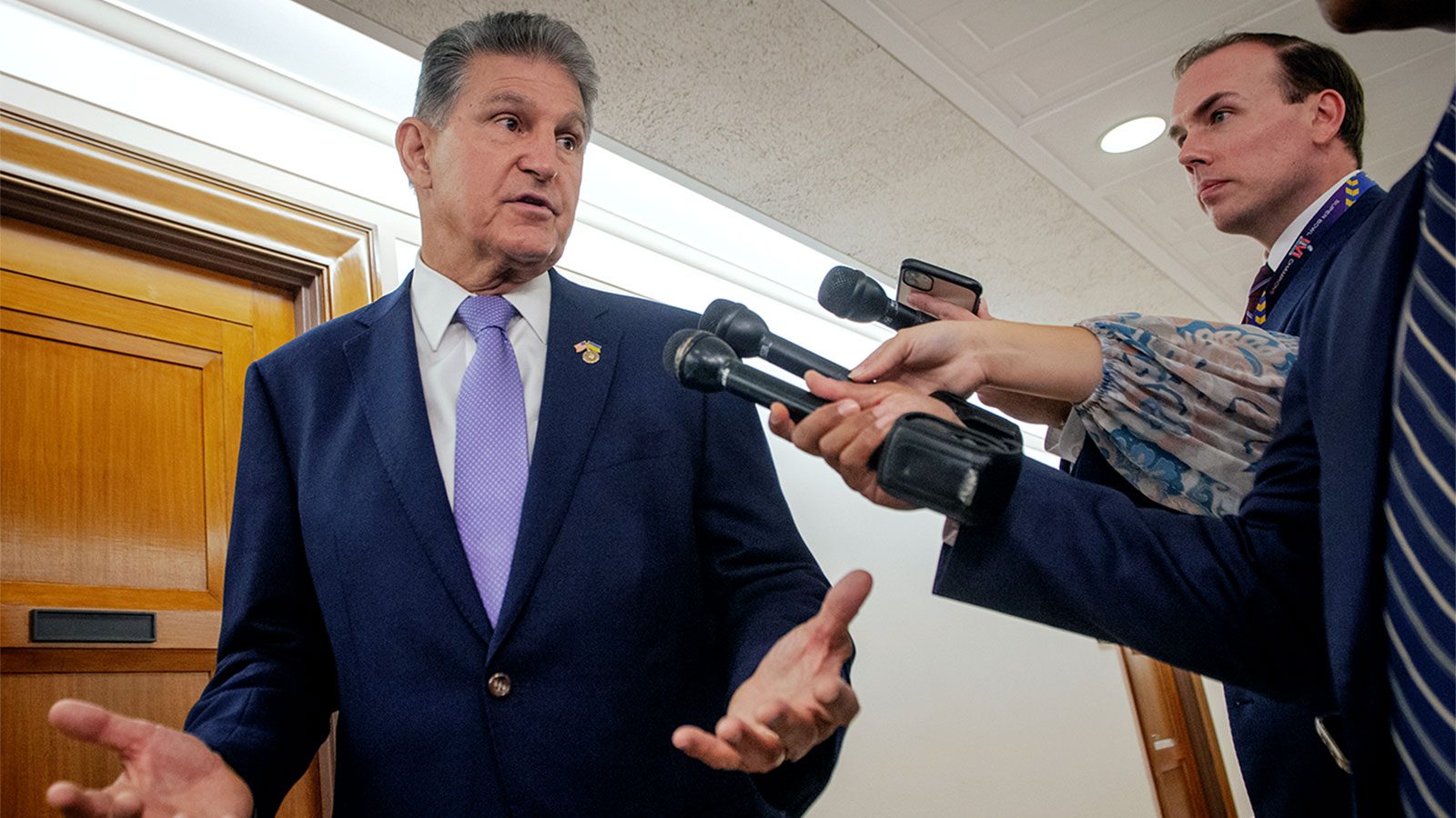 Reporters holding microphones out to Senator Joe Manchin, speaking with his hands raised in a half shrug