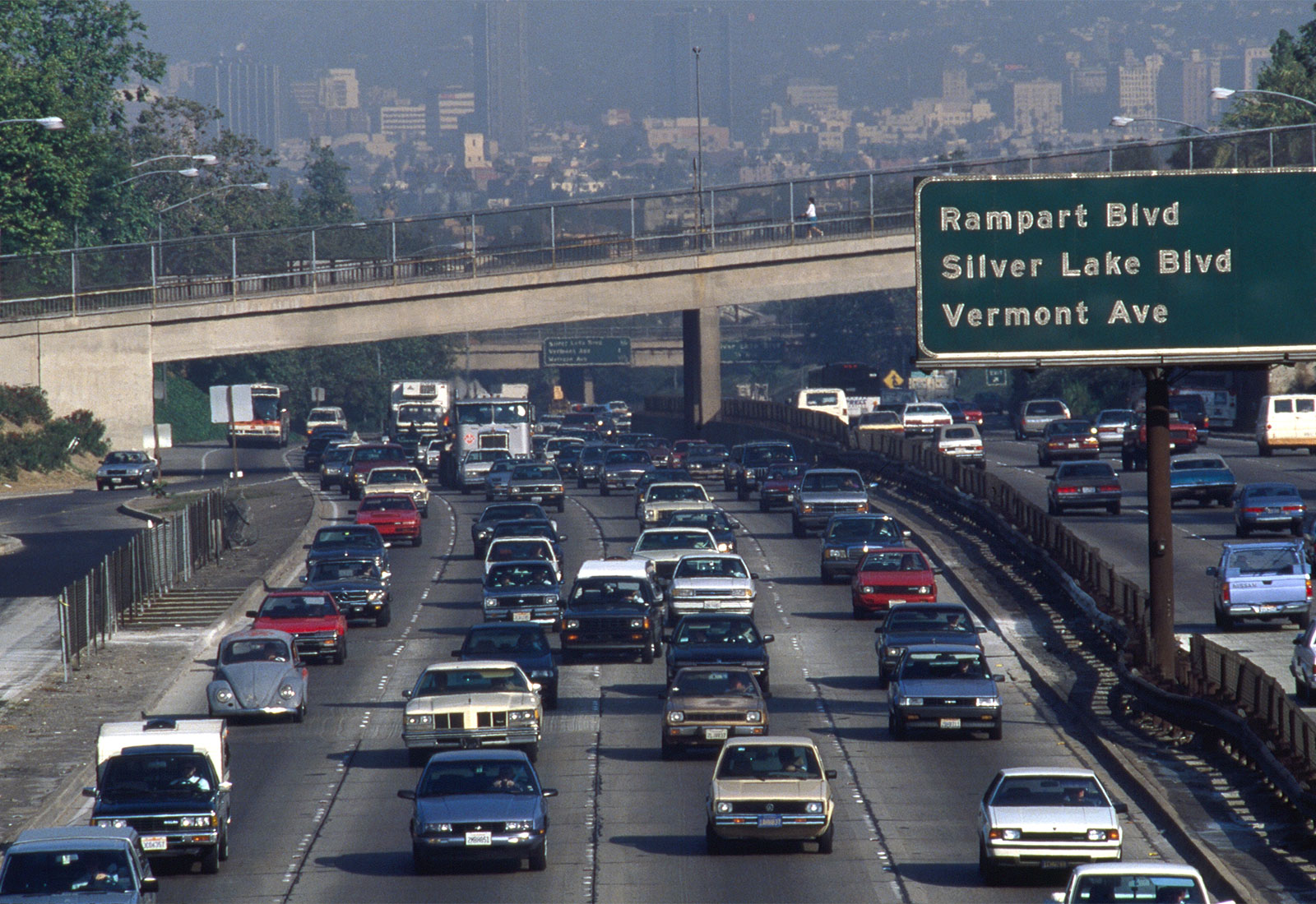 Freeway packed with cars on both sides with sign for exits in Los Angeles with bridge and buildings in distance