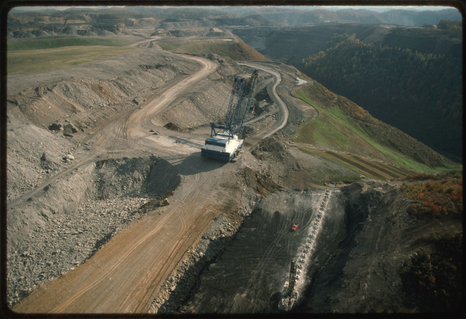 Aerial view of a mountaintop removal site with a large crane in the middle and tree covered mountains in the distance