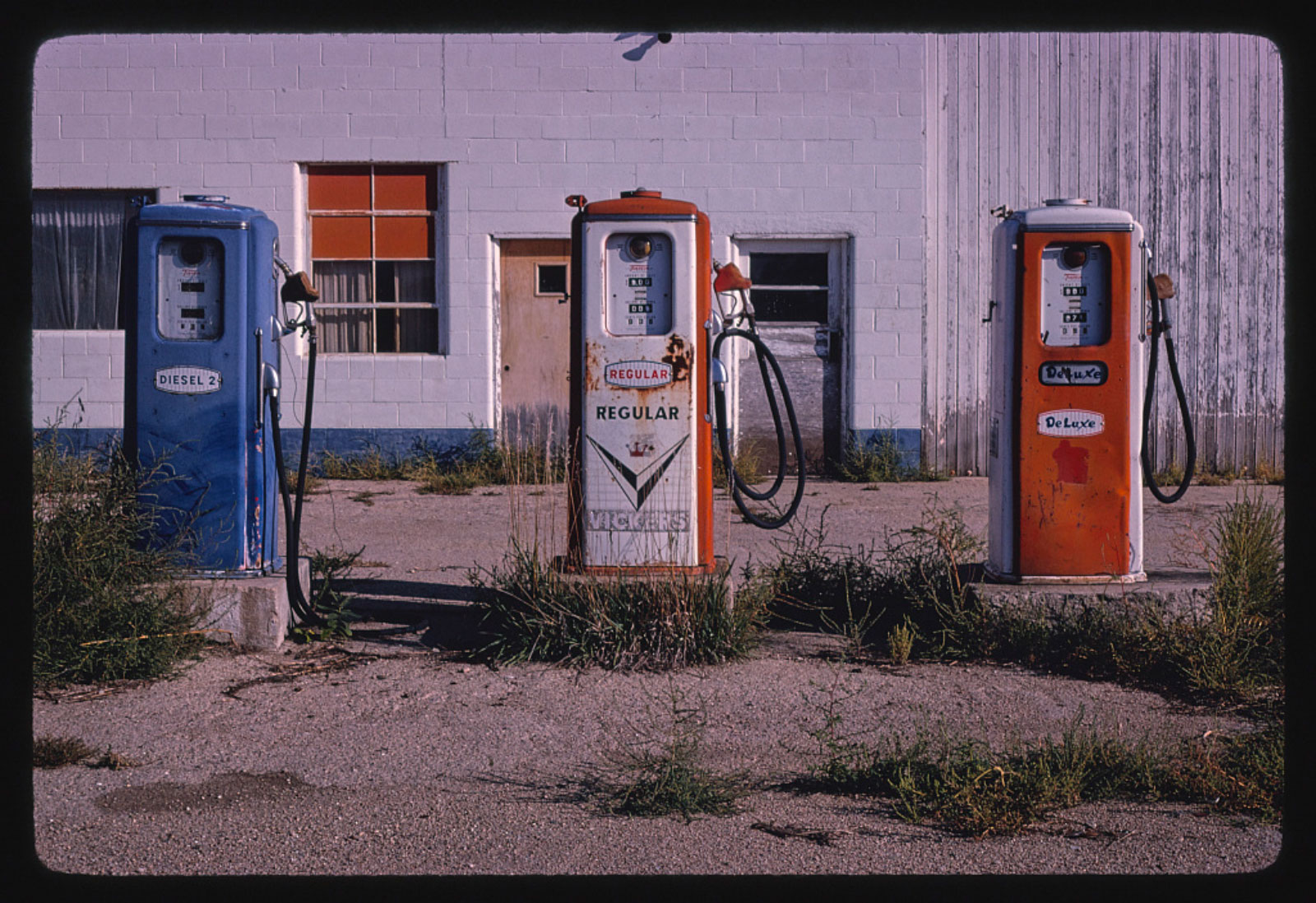 Photo circa 1980 of old rusted gas station pumps with a white brick and wood building in the background