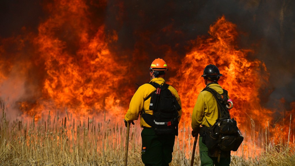 two firefighters stand looking at a huge red blaze