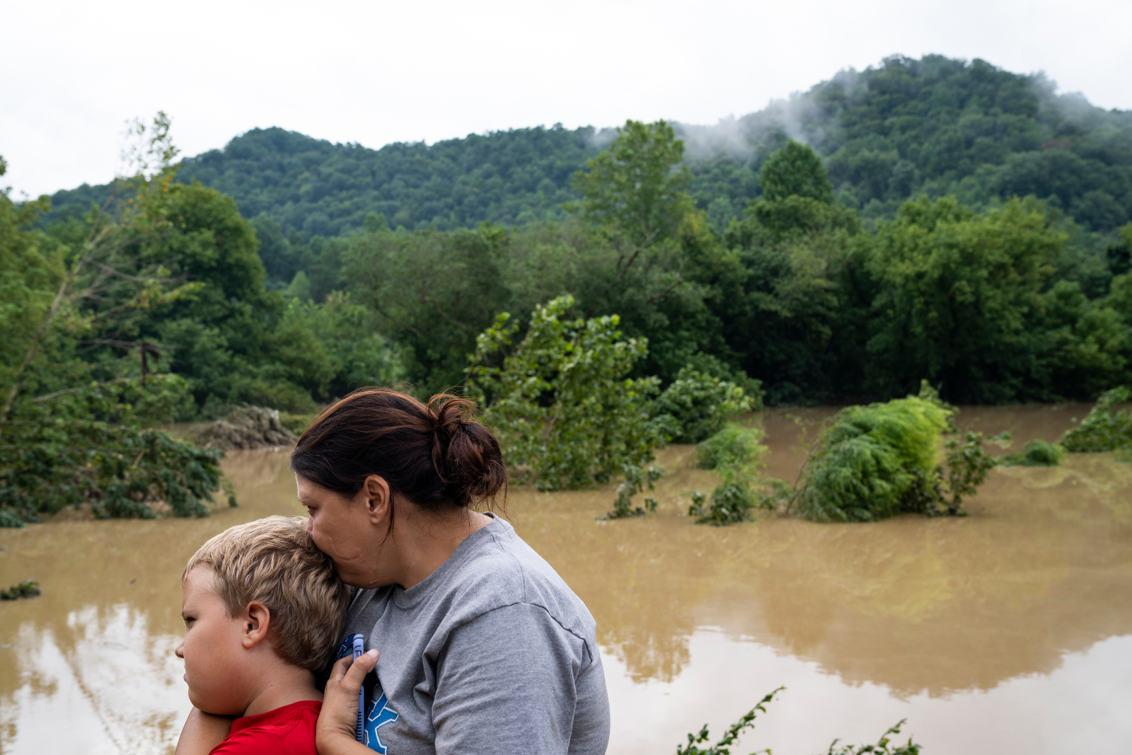 A women with brown hair holds a boy with blond hair. In the background, floodwaters are seen with debris and downed trees in front of a mountain landscape.