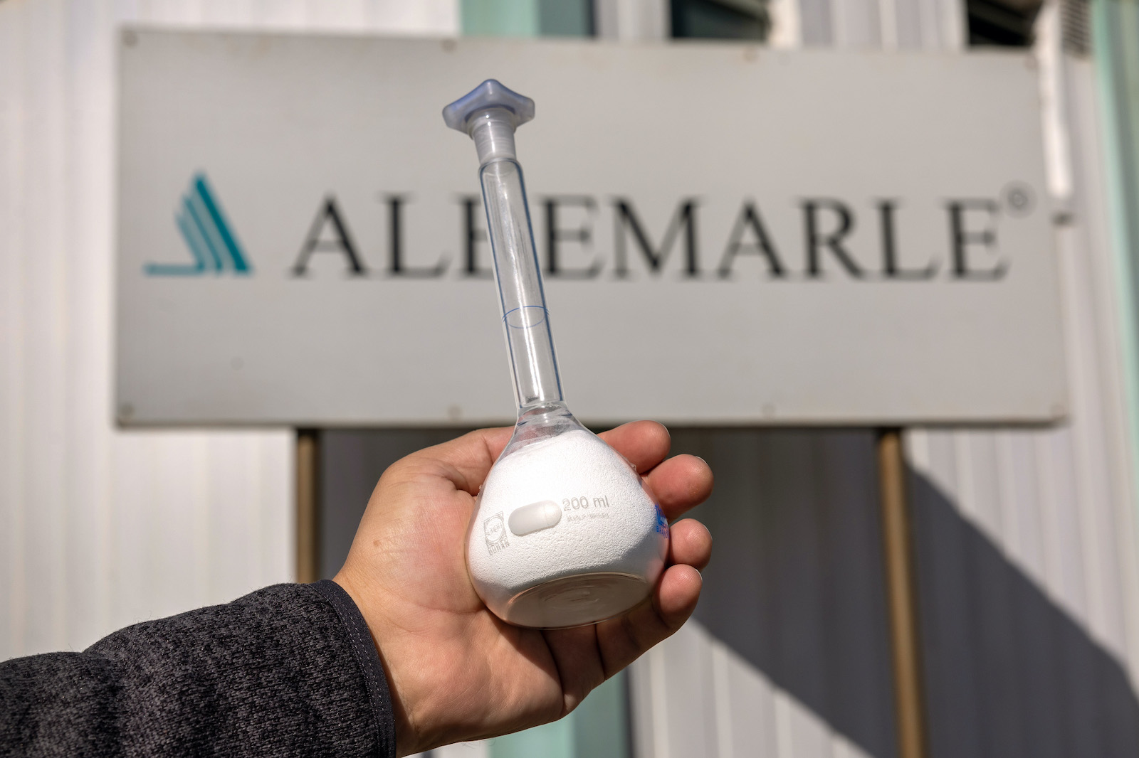 a hand holds a beaker of white powder in front of a sign that says albemarle