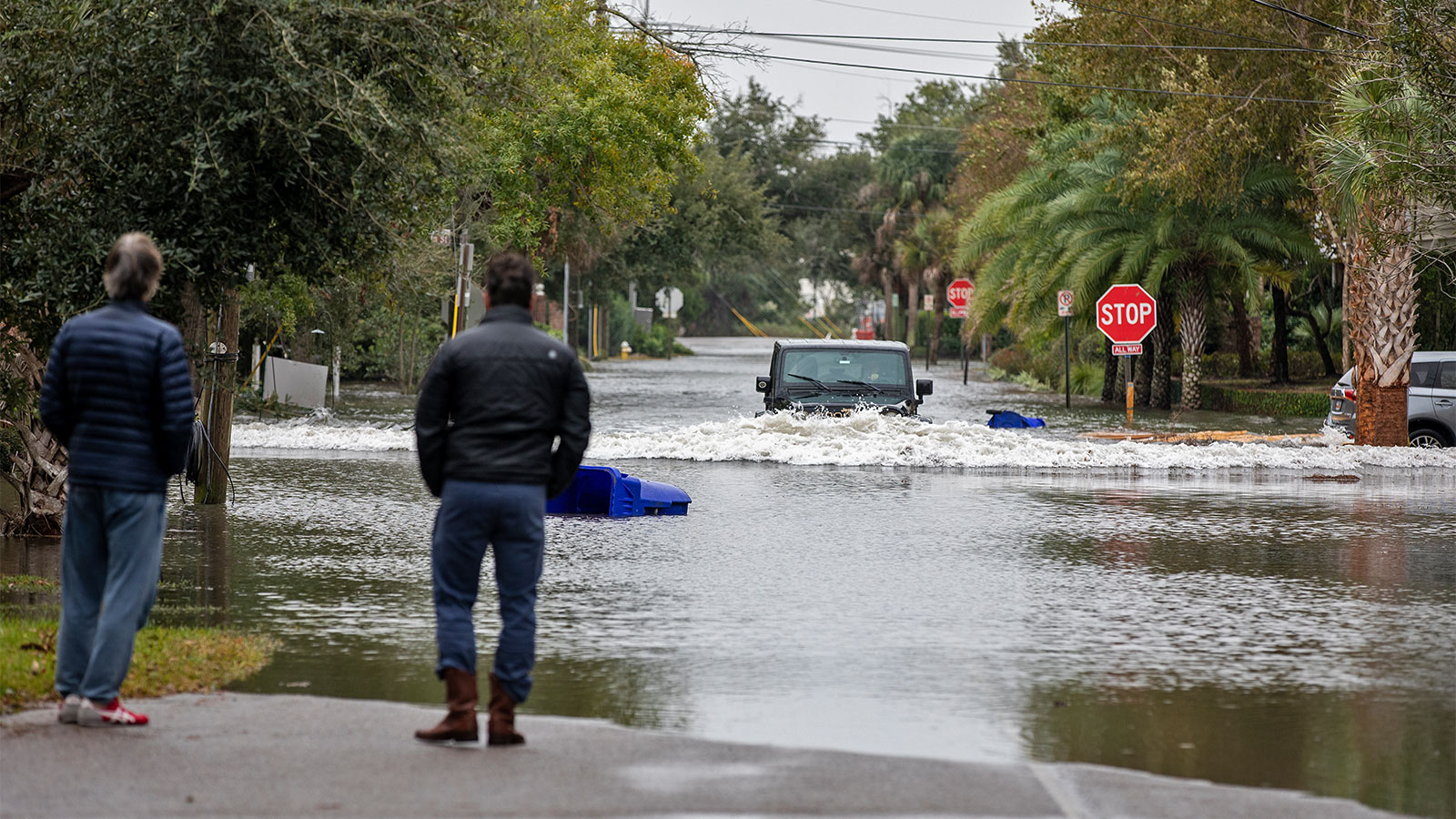 Two people looking down a flooded street at a Jeep driving through feet of water