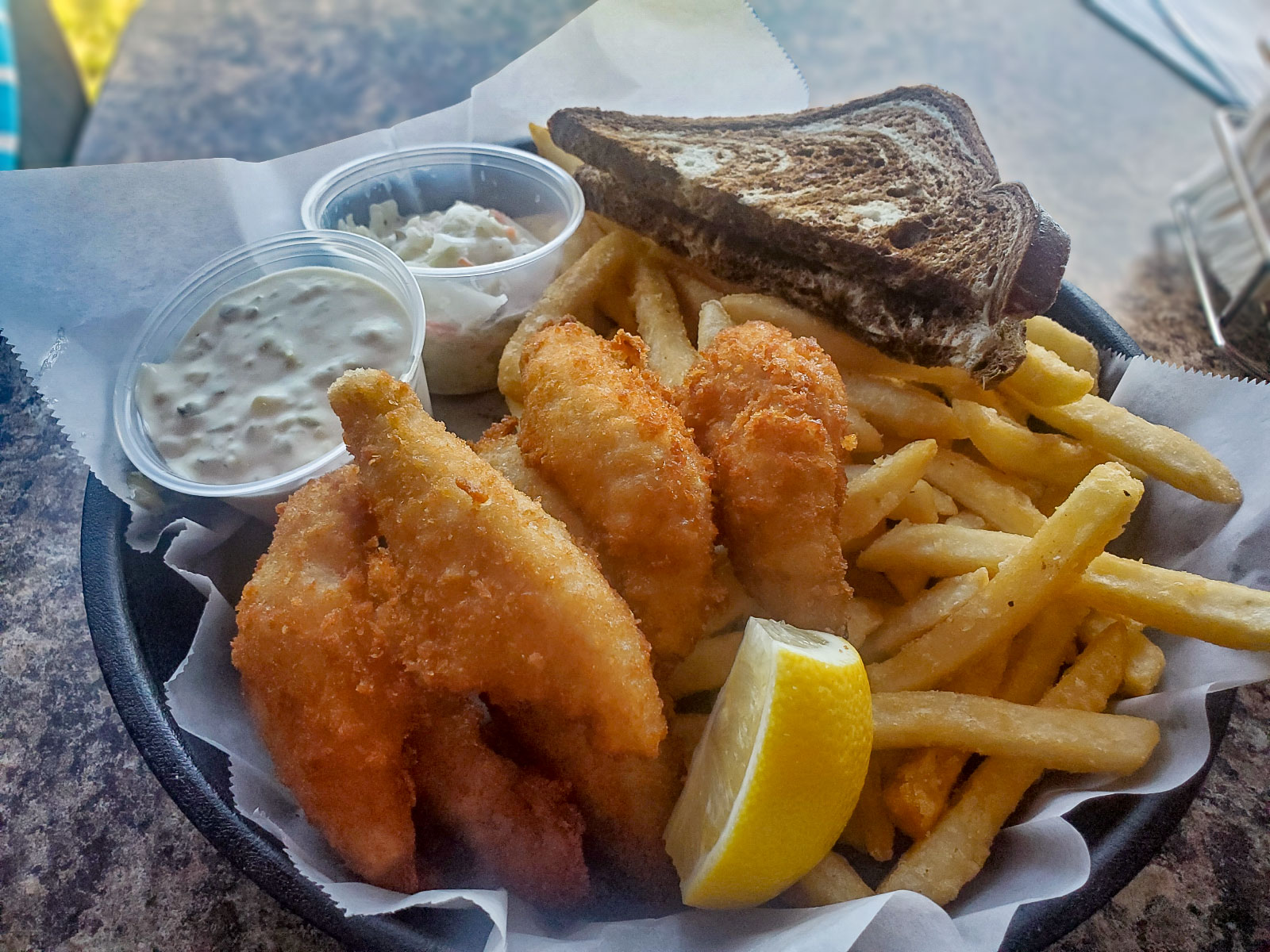 A plate of fried fish, lemon slice, french fries, white sauce, and brown and white rye bread