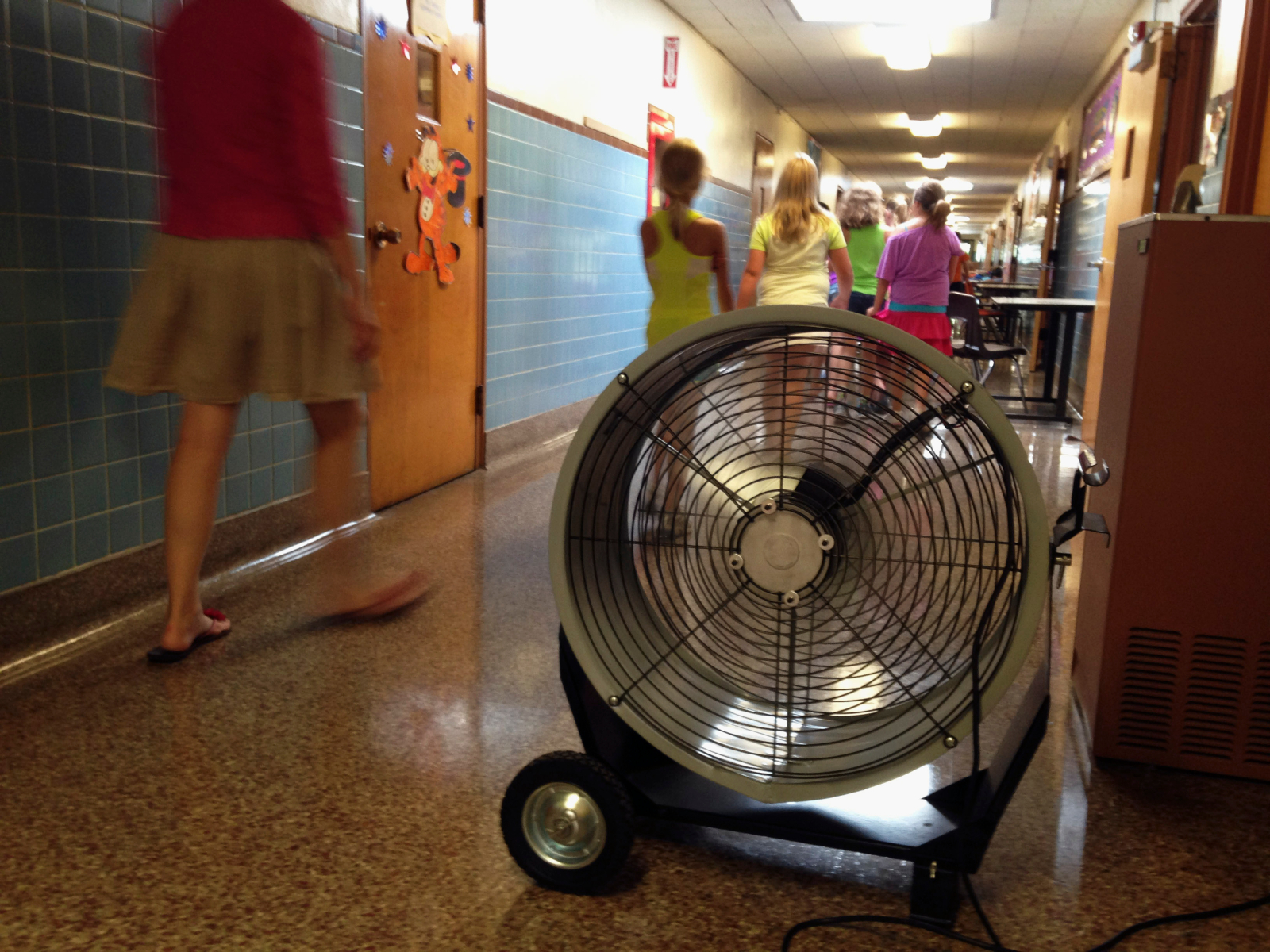 Three school children and a teacher walk through a hallway with blue tile on the wall. A large floor fan is seen in the middle of the hallway.
