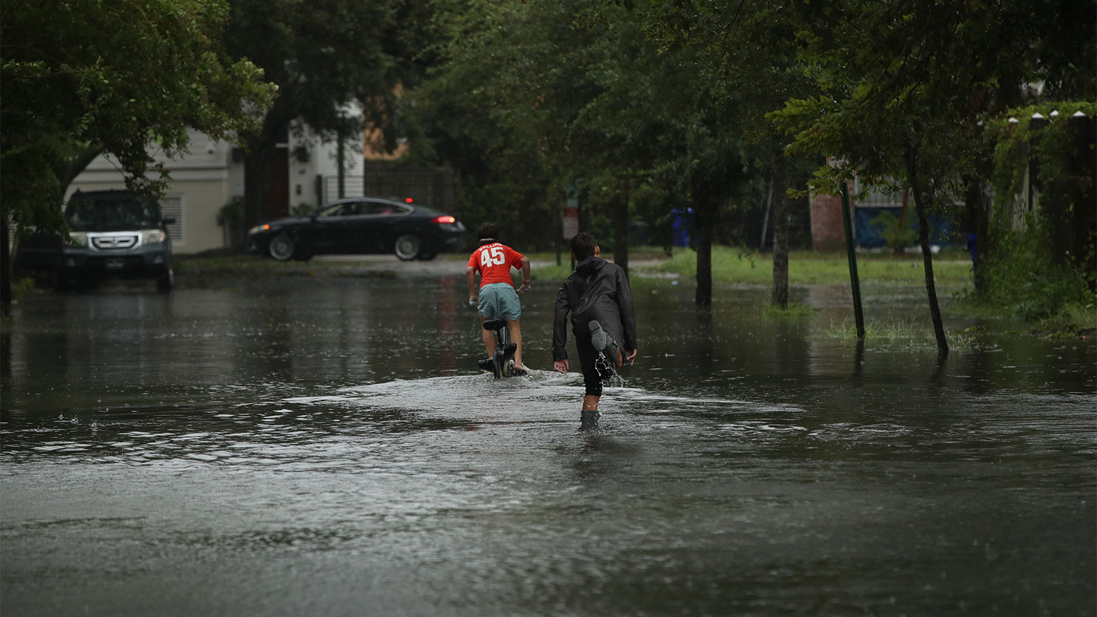 Two youths on a flooded street; one on a bike and one skateboarding; houses and trees in the distance