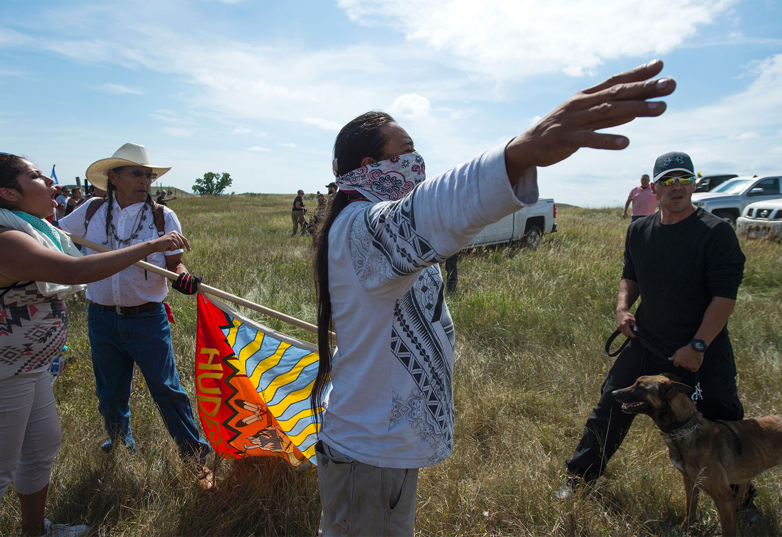 Indigenous people raise their arms and a flag at a security guard holding a dog by the leash at the Dakota Access Pipeline site