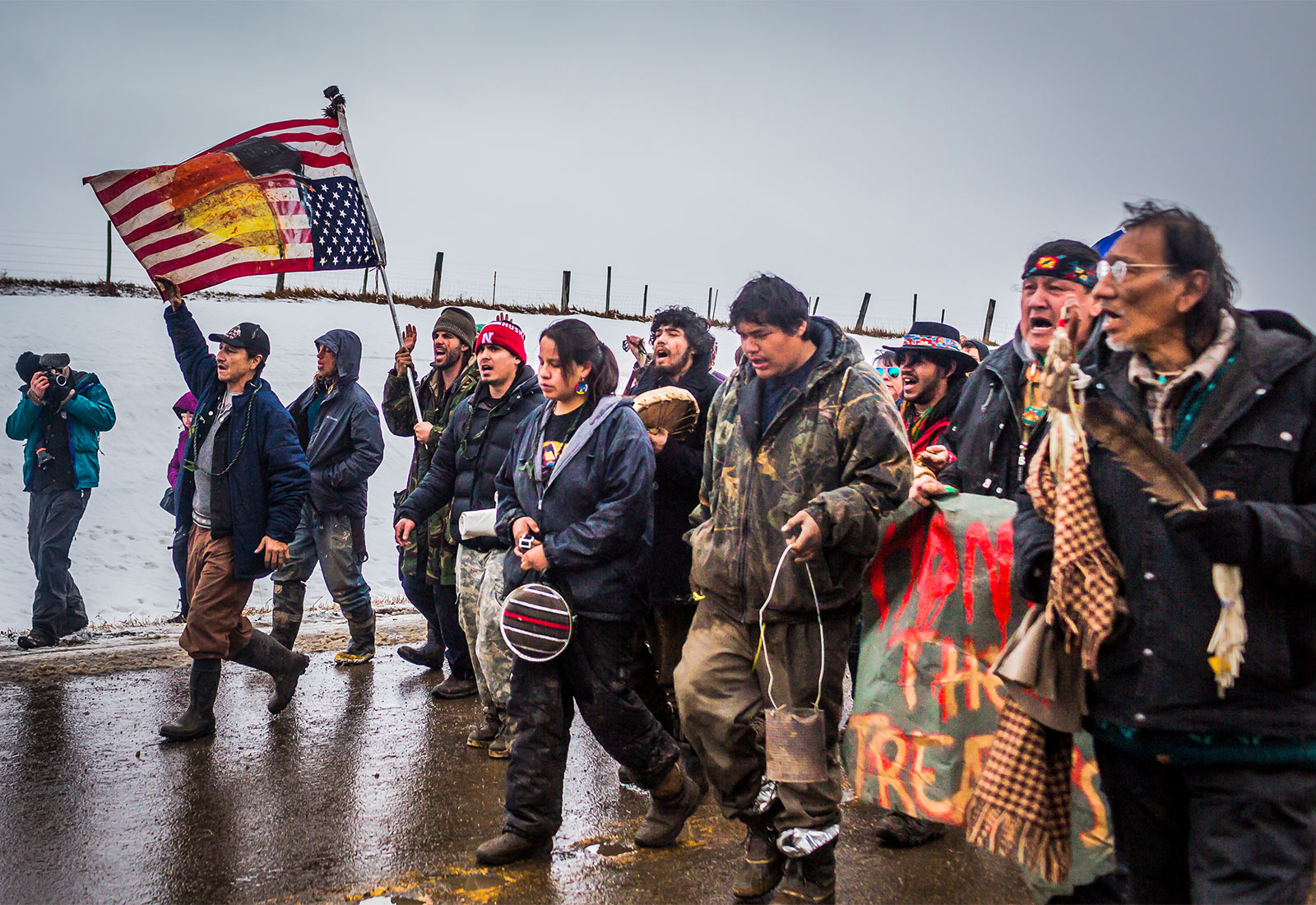 A large group of Indigenous people march and protest on a wet road on a snowy field