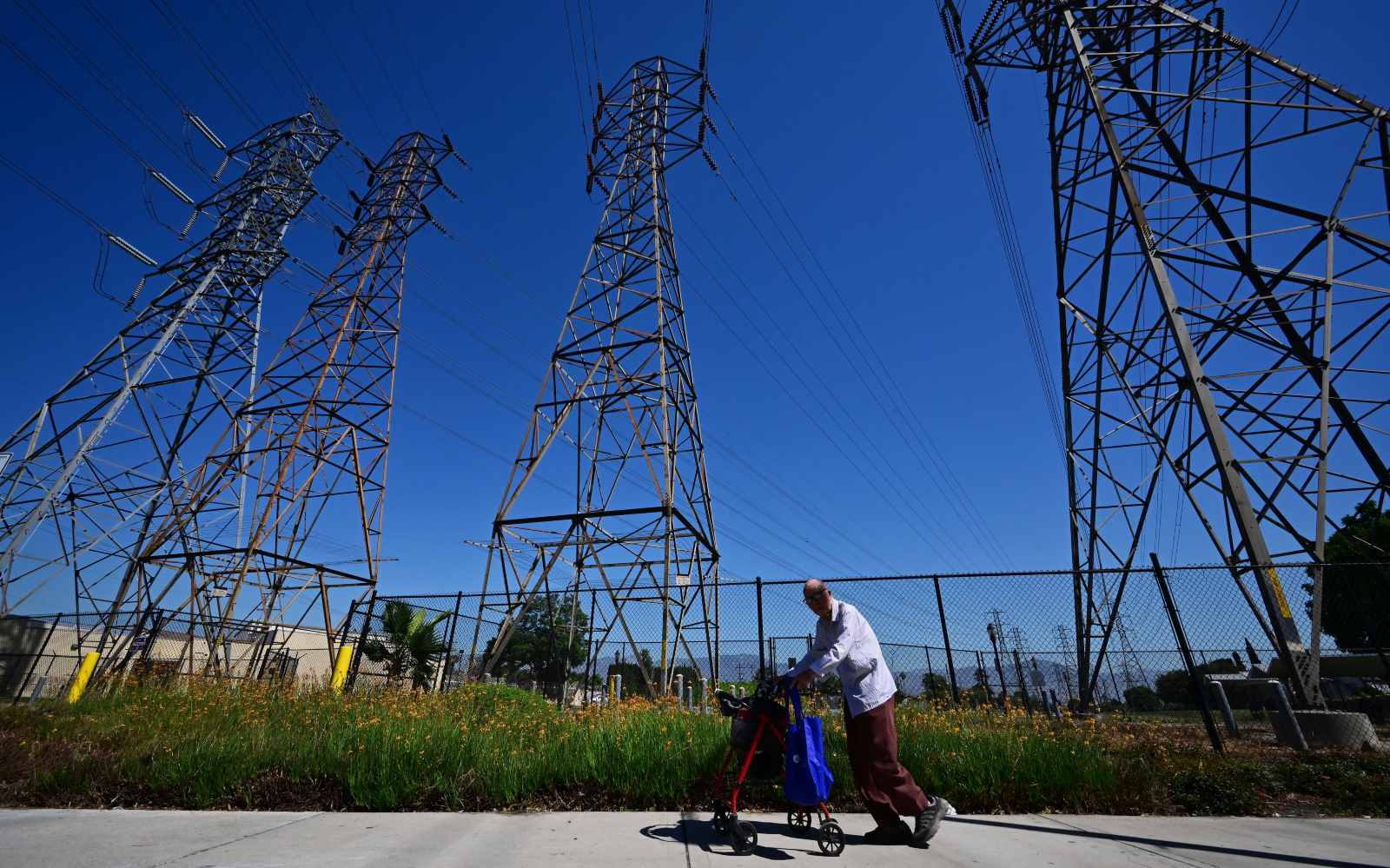 An elderly man with a walker stands in front of large power line on a sunny, clear skies day.