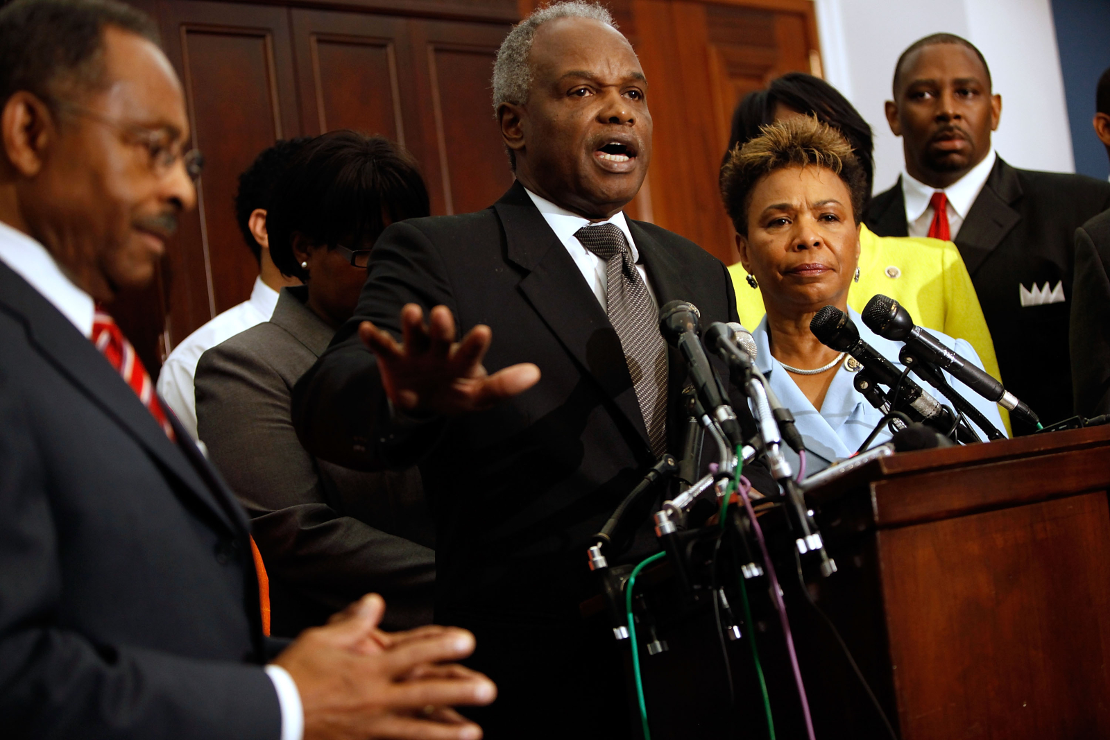 A Black man with short, gray hair speaks into a group of microphones on a podium. He is surrounded by other Black people wearing formal suits and clothing.