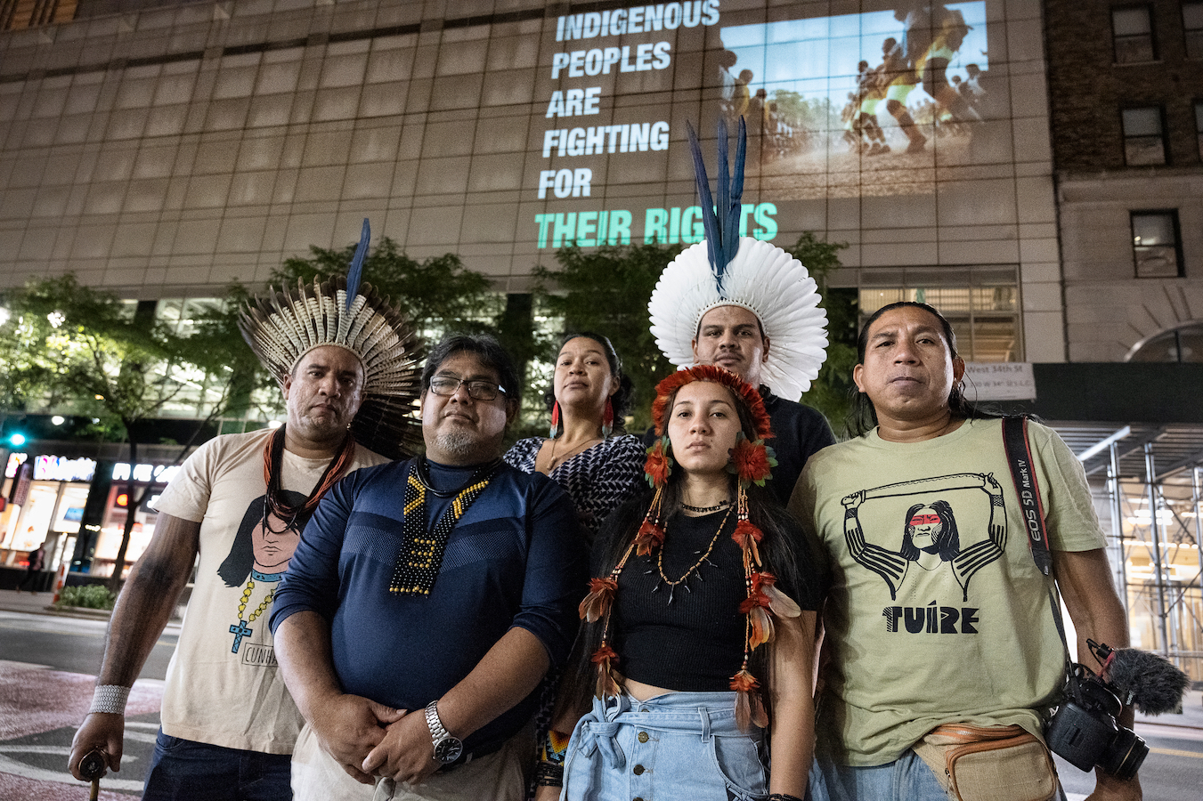 six Indigenous people stand in front of a projected message on a building that reads 