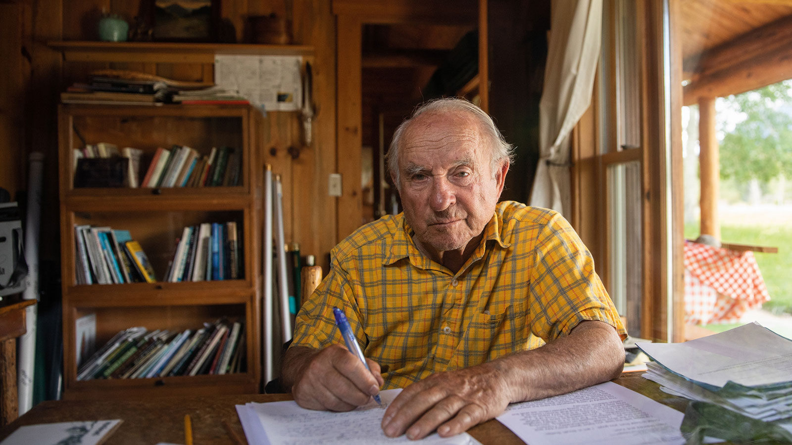 Portrait of Yvon Chouinard, the founder of Patagonia, seated at a desk writing on a piece of paper and looking at the camera