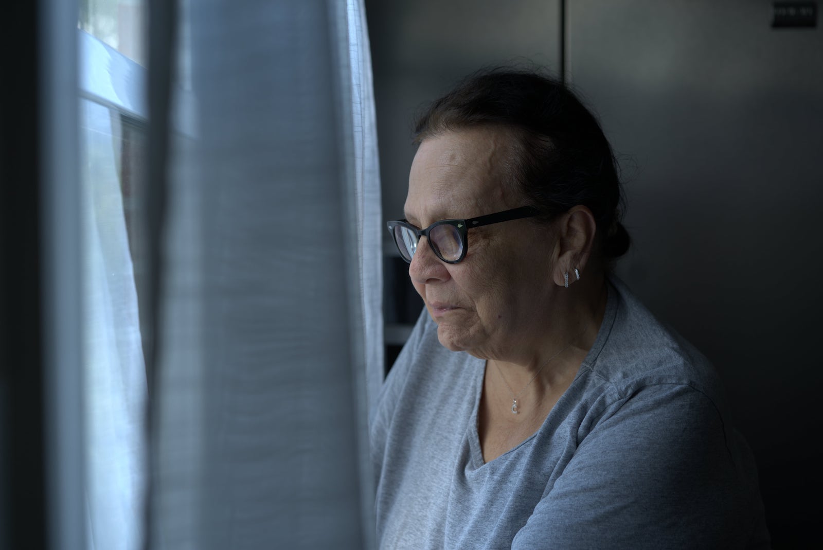 an older woman with glasses looks through a curtain