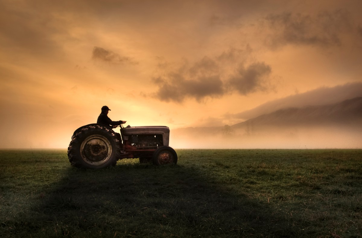 A tractor sits in a field of green grass. it is dark outside and the sun is rising in the background.