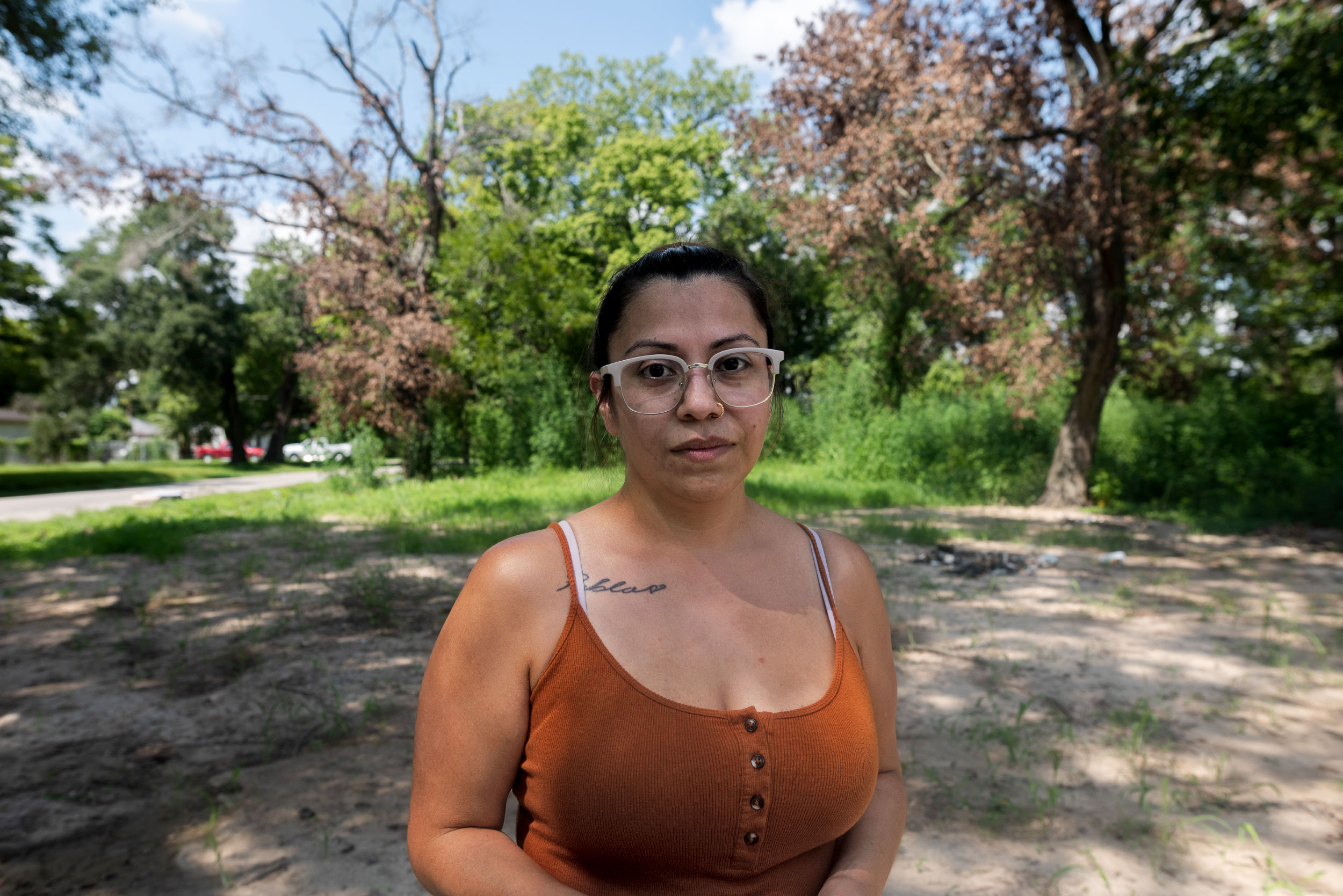 A woman with glasses and an orange tank top centered. In the background, trees and dead grass