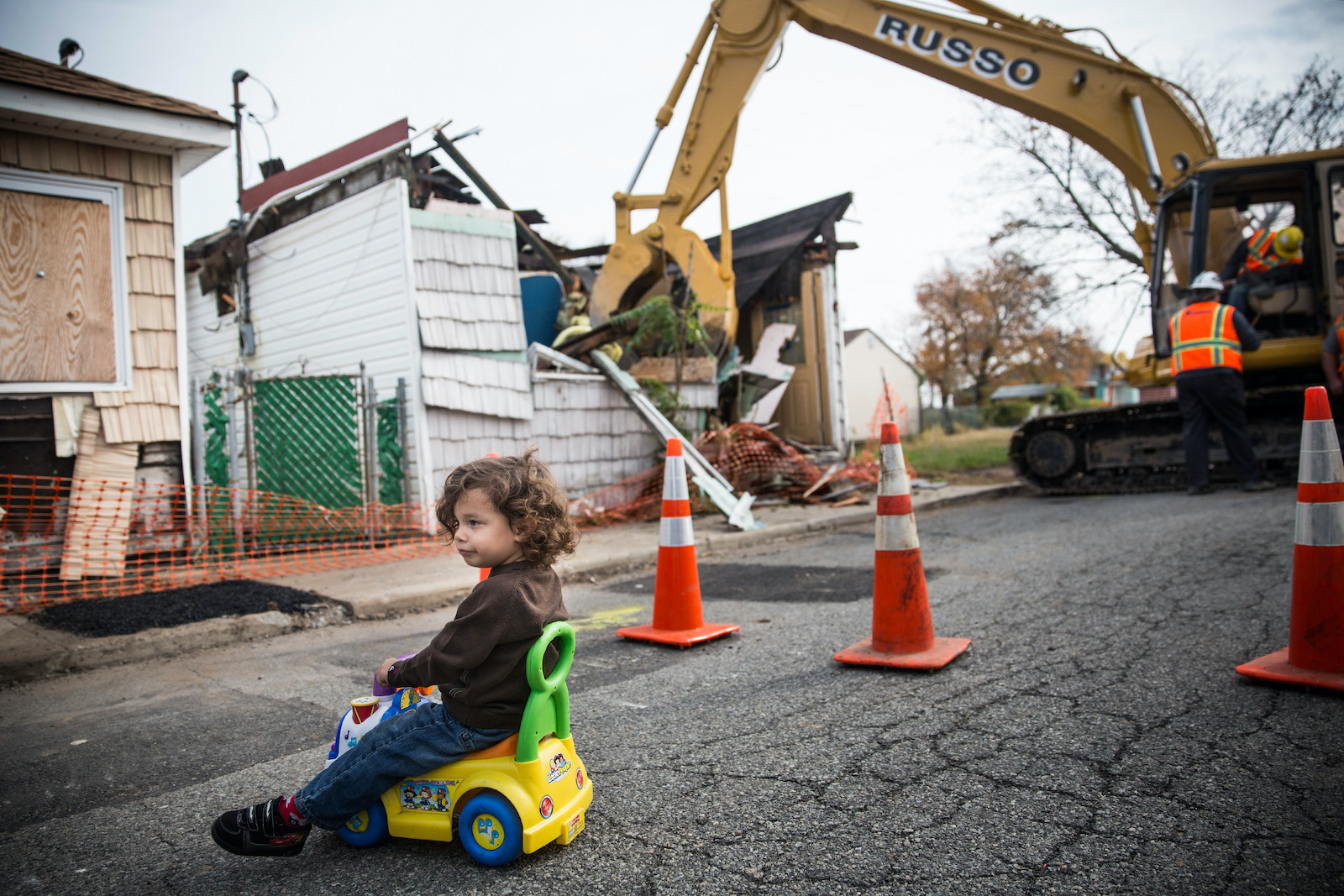 a child in a toy car plays on a street. IN the background, an excavator takes apart a damaged house