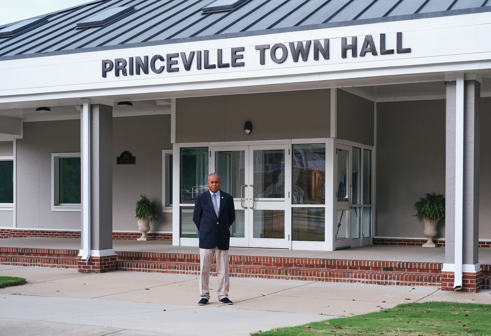 a man in a suit stands in front of a white building with the words Princeville Town Hall marked on it