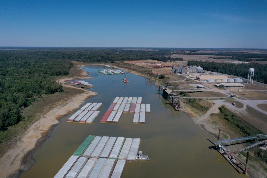 An aerial image shows various container ships sit in the Mississippi River, to the left is green vegetation and to the right is farm equipment