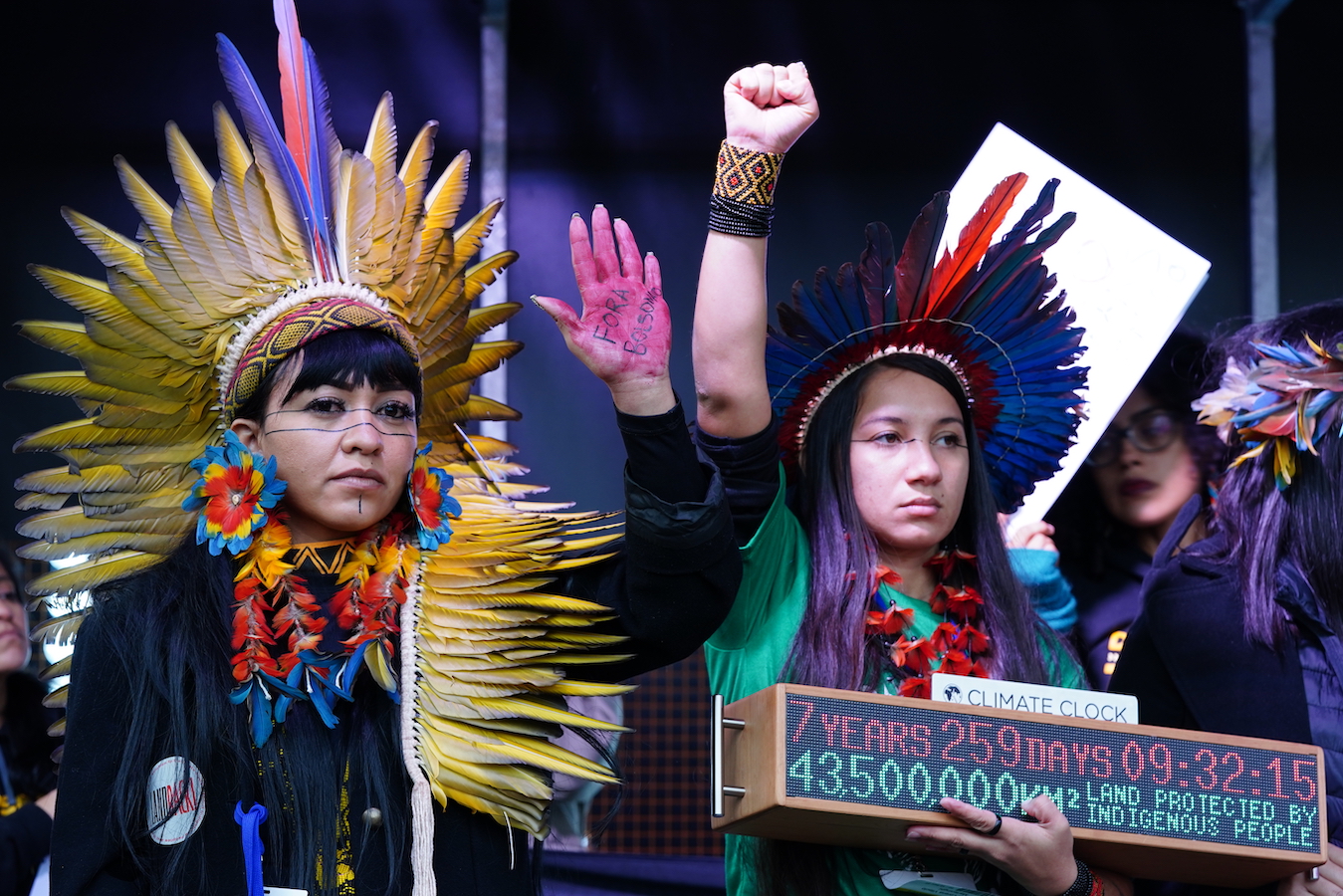 two Indigenous people wearing feathered headdresses raise their hands