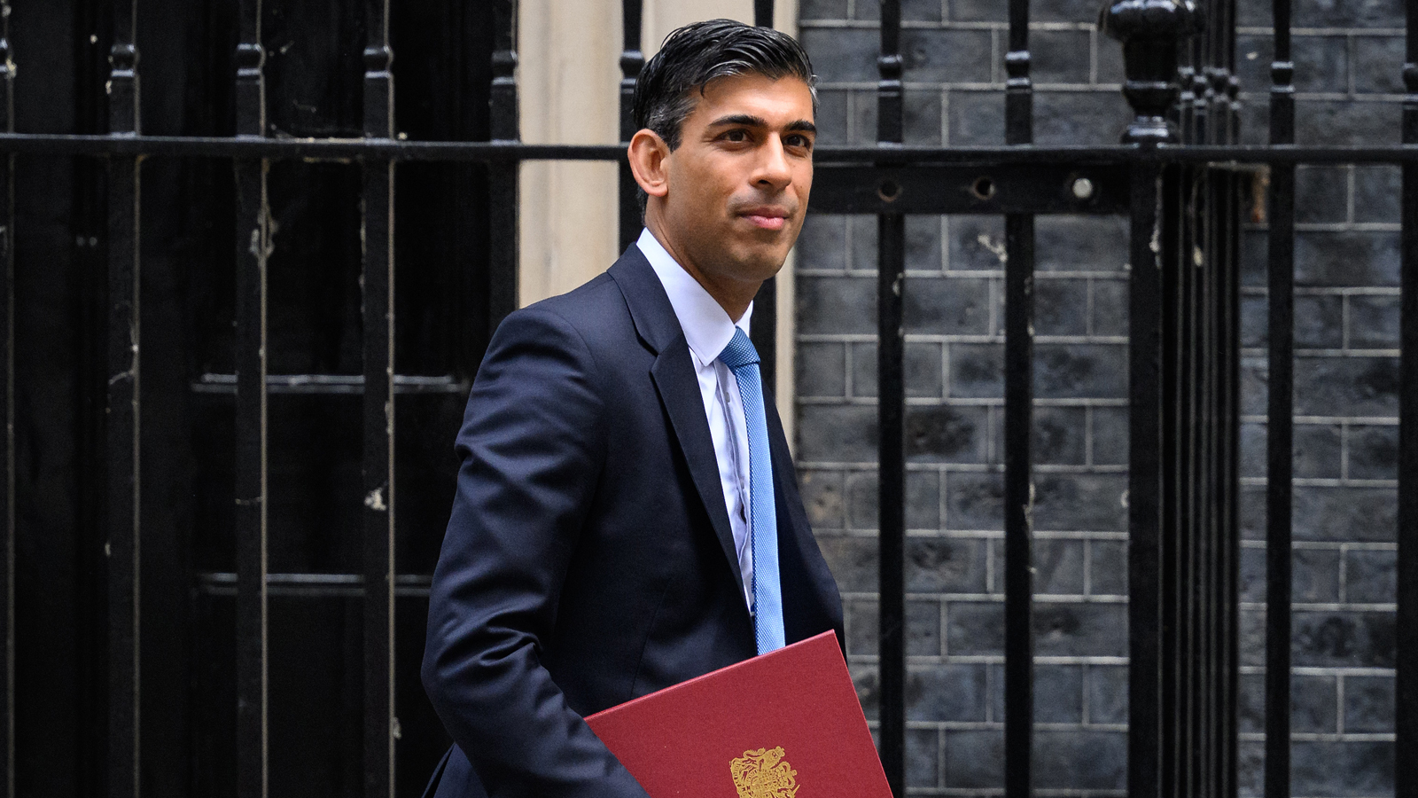 new UK prime minister rishi sunak walkins in front of a fence holding documents and wearing a suit