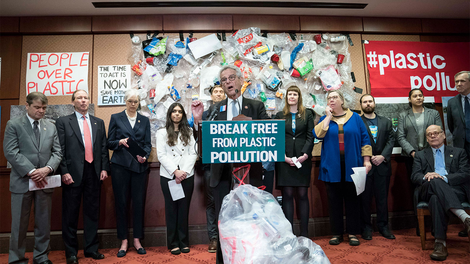 Rep. Alan Lowenthal speaking at a podium with a sign reading "break free from plastic pollution"; a group of people stands behind him in front of a wall with plastic trash attached to it
