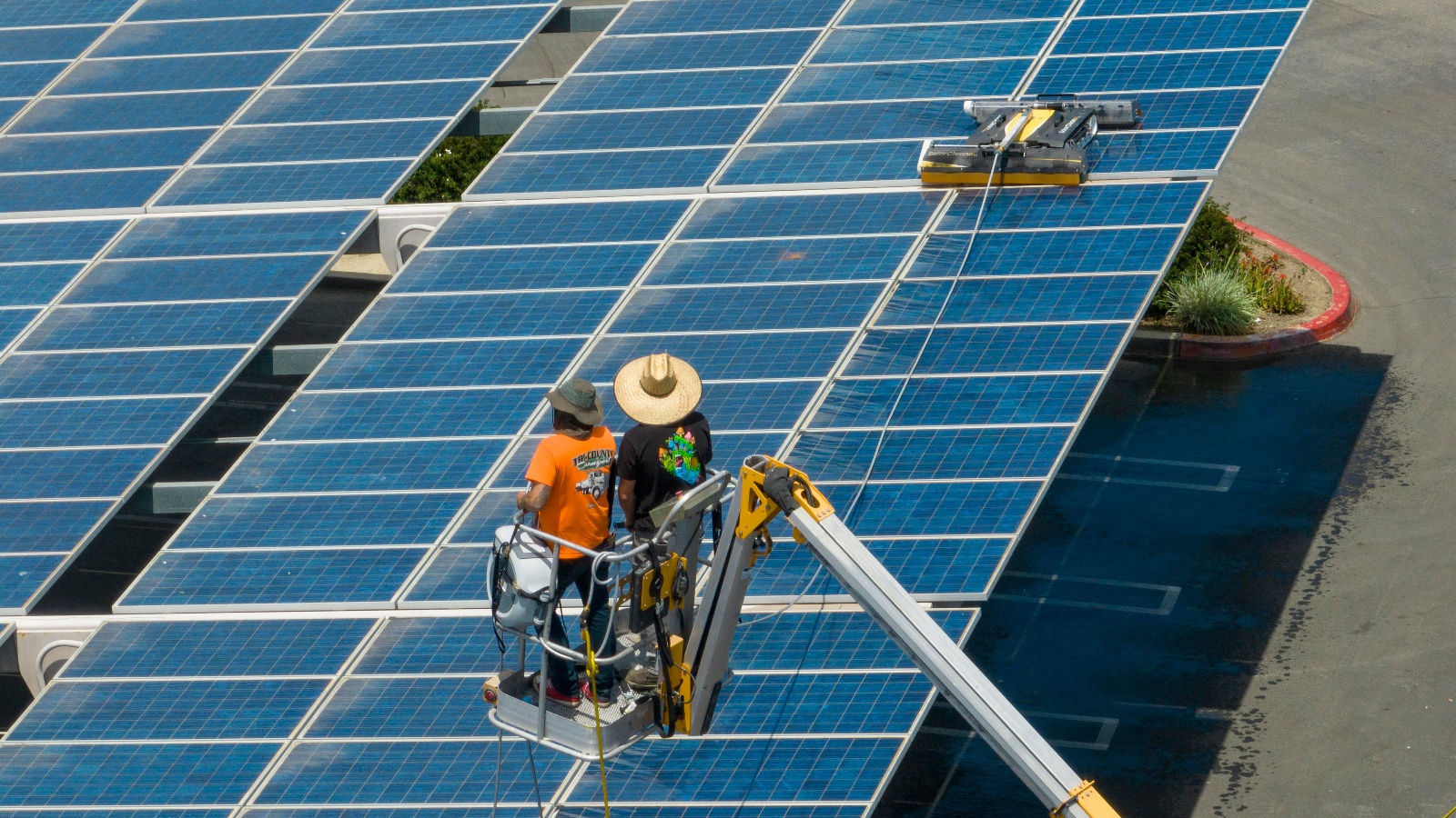 Workers on an elevated crane watch a cleaning robot wash large solar panels