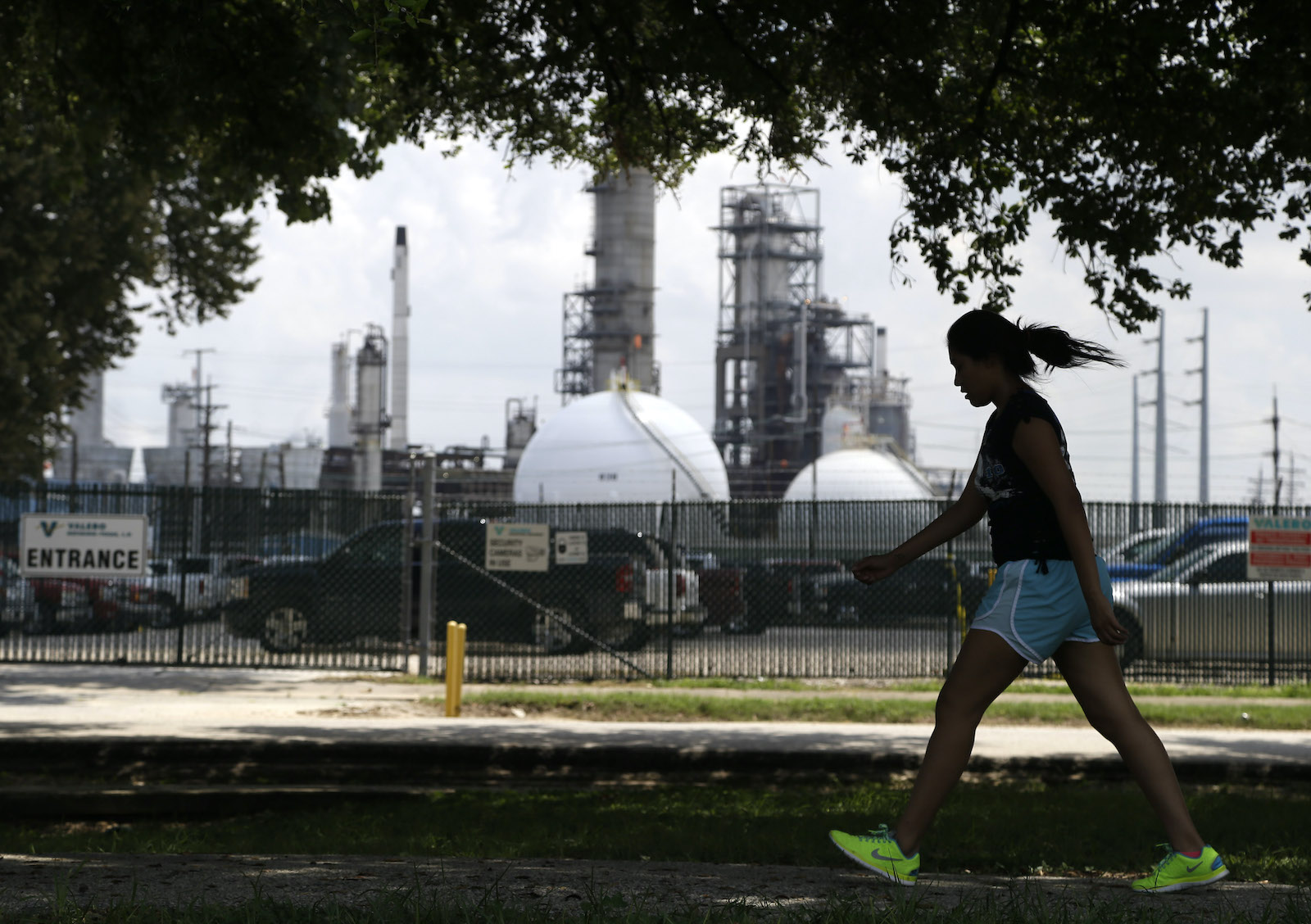 a girl walks in a park with an oil refinery in the background