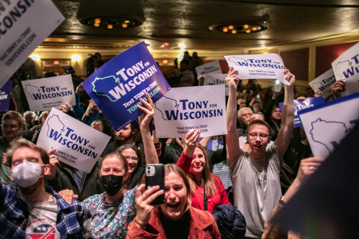 Varied people, mostly white, hold blue and white signs that read "Tony for Wisconsin" and are cheering and smiling inside a packed room.