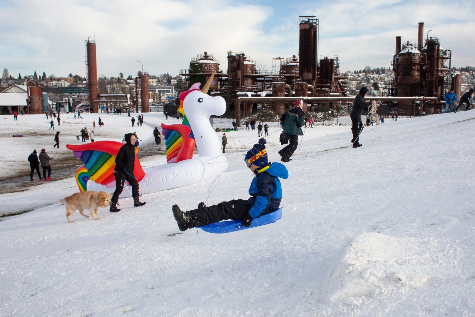 Kids sled down a snowy hill near an industrial factory.