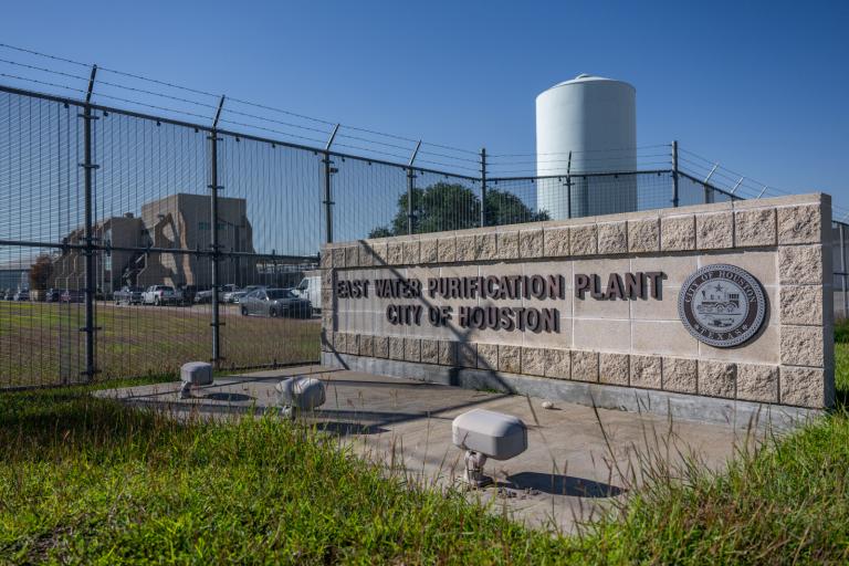 A sign that reads "East Water Purification Plant, City of Houston" sits outside of a fenced in, blue and gray industrial building