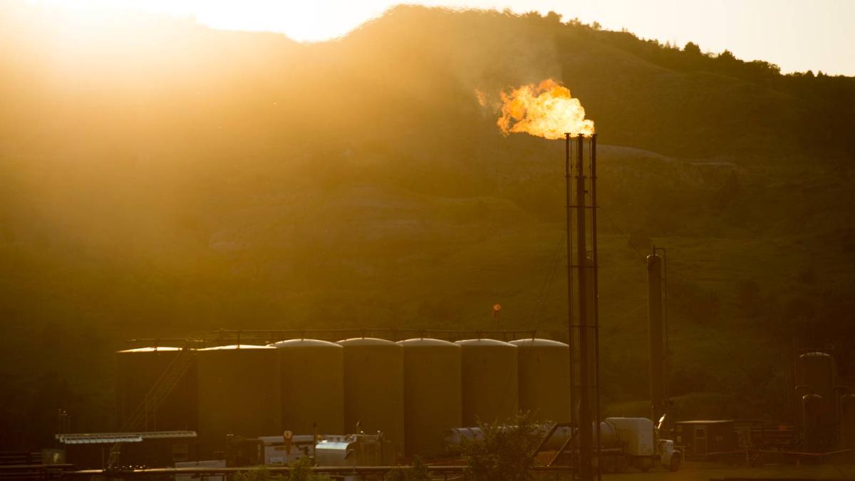A gas well sits on the Fort Berthold Indian Reservation as the sun sets behind a ridge line. A tall metal structure is "flaring", or burning off natural gas, and a bright orange flame is spewing from the top of the structure.