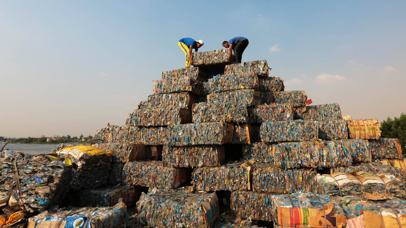 Two people lift blocks of plastic on top of a pyramid made of trash