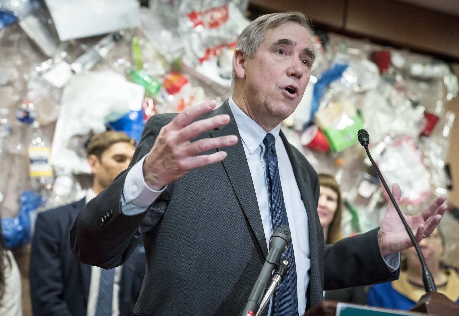 Sen. Jeff Merkley speaks at a podium with plastic trash in the background