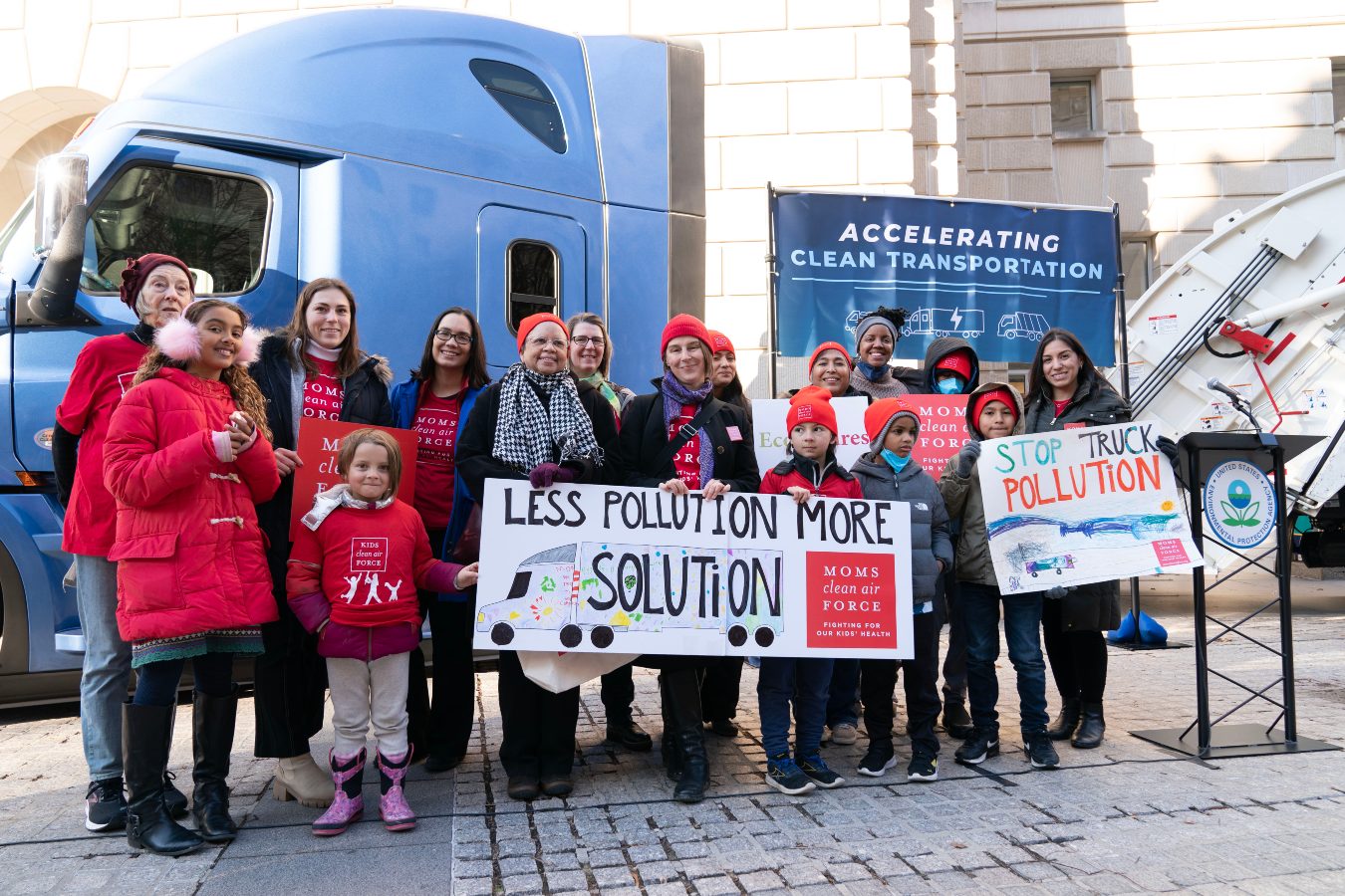 A group of women and children dressed for cold weather hold signs against truck pollution in front of a stationary blue-colored semi truck on December 20, 2022.