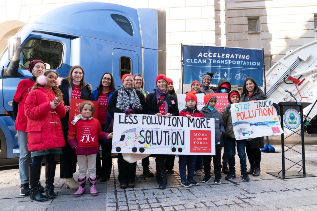 A group of women and children dressed for cold weather hold signs against truck pollution in front of a stationary blue-colored semi truck on December 20, 2022.
