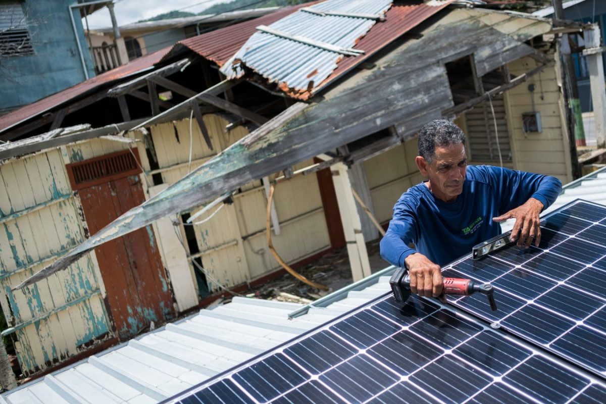 A man in a blue shirt holding a tool installs a solar panel on the roof of a home in Puerto Rico