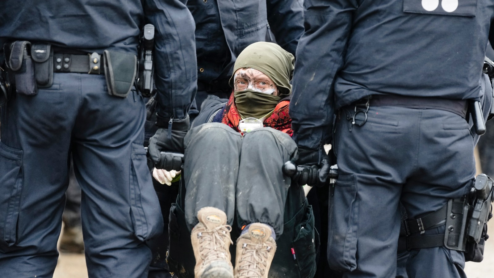 Police in dark suits carry a protestor away from the coal mine. The protestor has dirt on their face and looks slightly off camera.