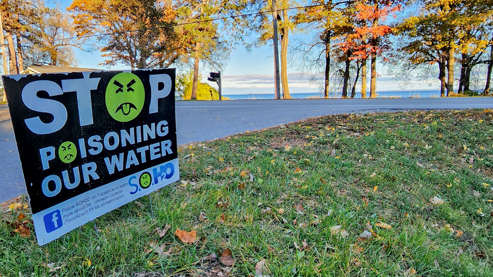 Low angle view of a sign reading "stop poisoning our water", with autumnal trees and a body of water behind it