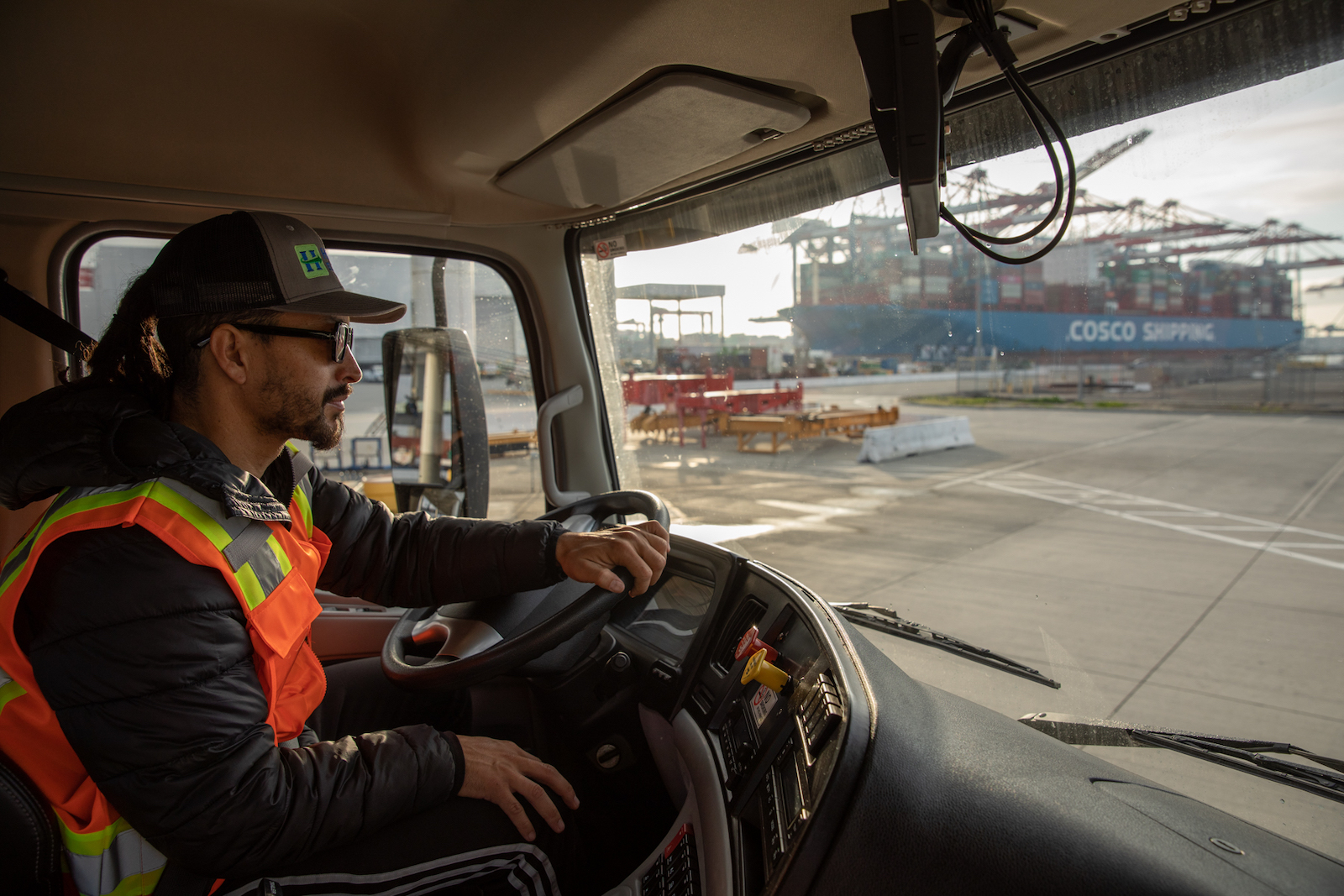 a man in an orange vest and trucker hat sits inside the cab of a large truck driving near shipping containers