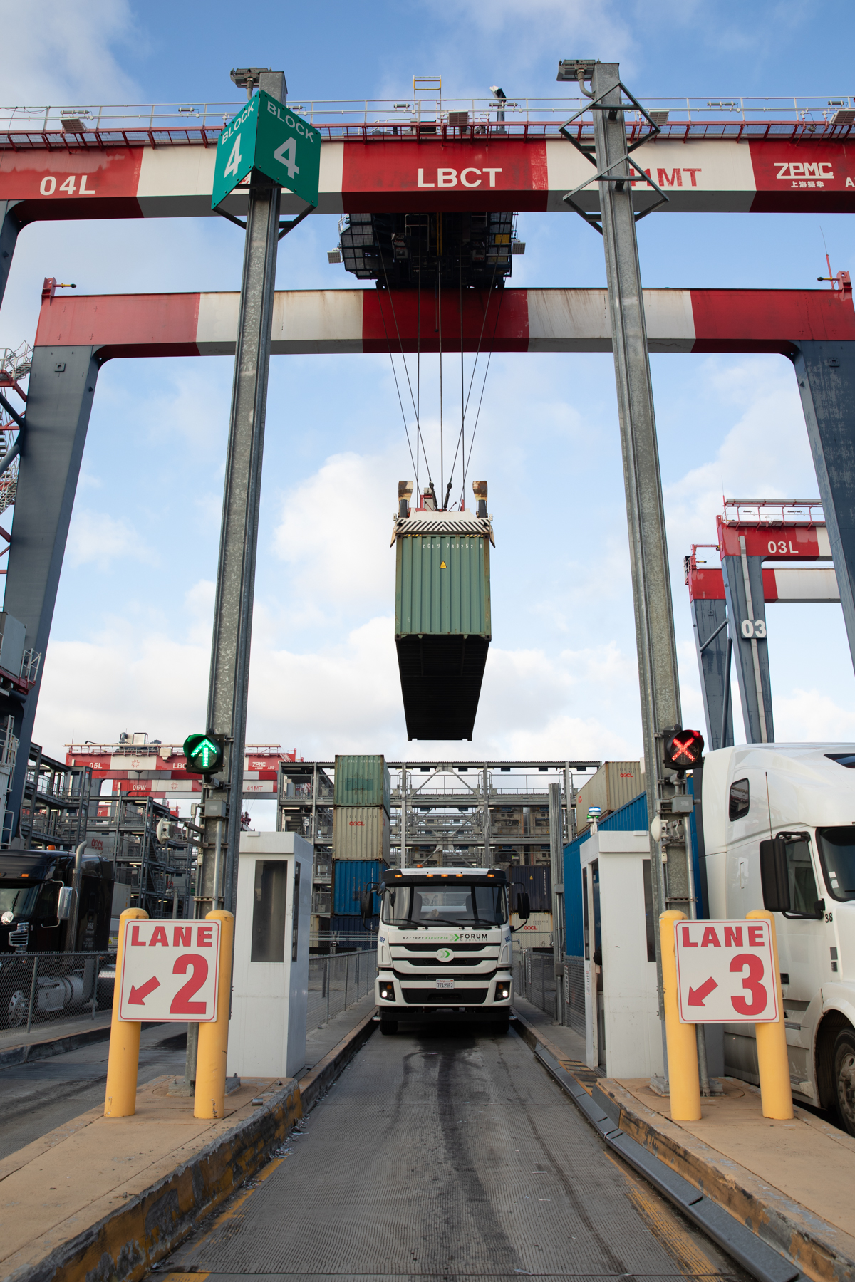 a green cargo container is lifted above a large truck underneath a red and white metal bar