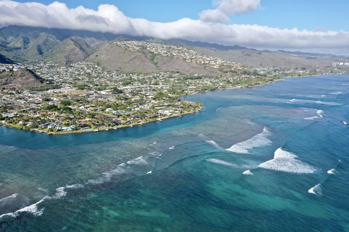 An aerial view of the Hawaii coastline with blue-green water lapping at the mountainous island.