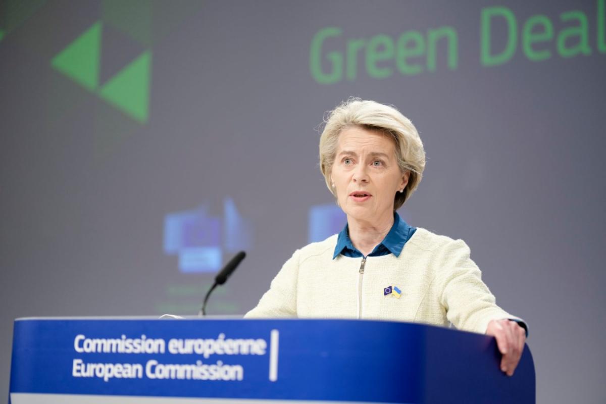 A woman in white zip-up top stands at a blue podium with the words "Green Deal" in the background.