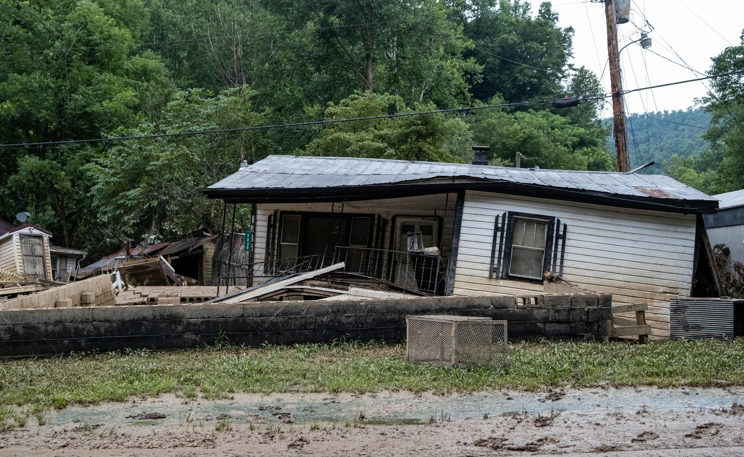 A damaged single story house with its front porch bent lies in a forested area in Eastern Kentucky after flooding in July 2022